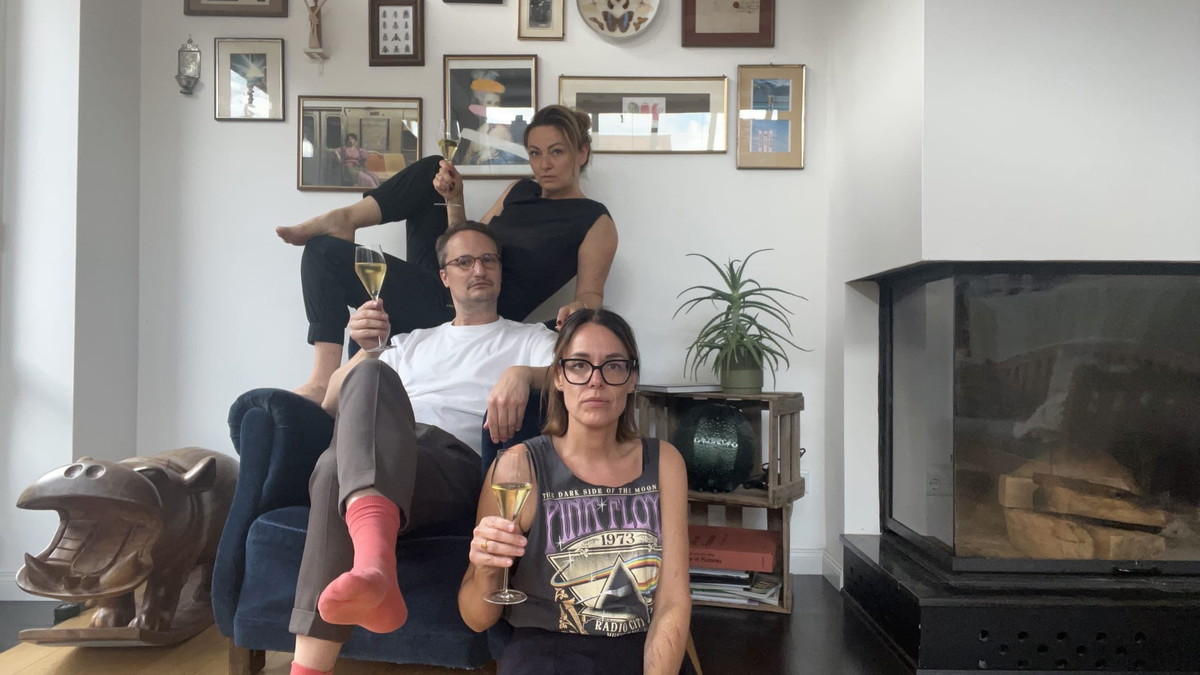 Dr. B, Dr. D und Kathrin M. vom Literaturhaus sitzen mit einem Glas Wein vor einer Wand mit Bildern