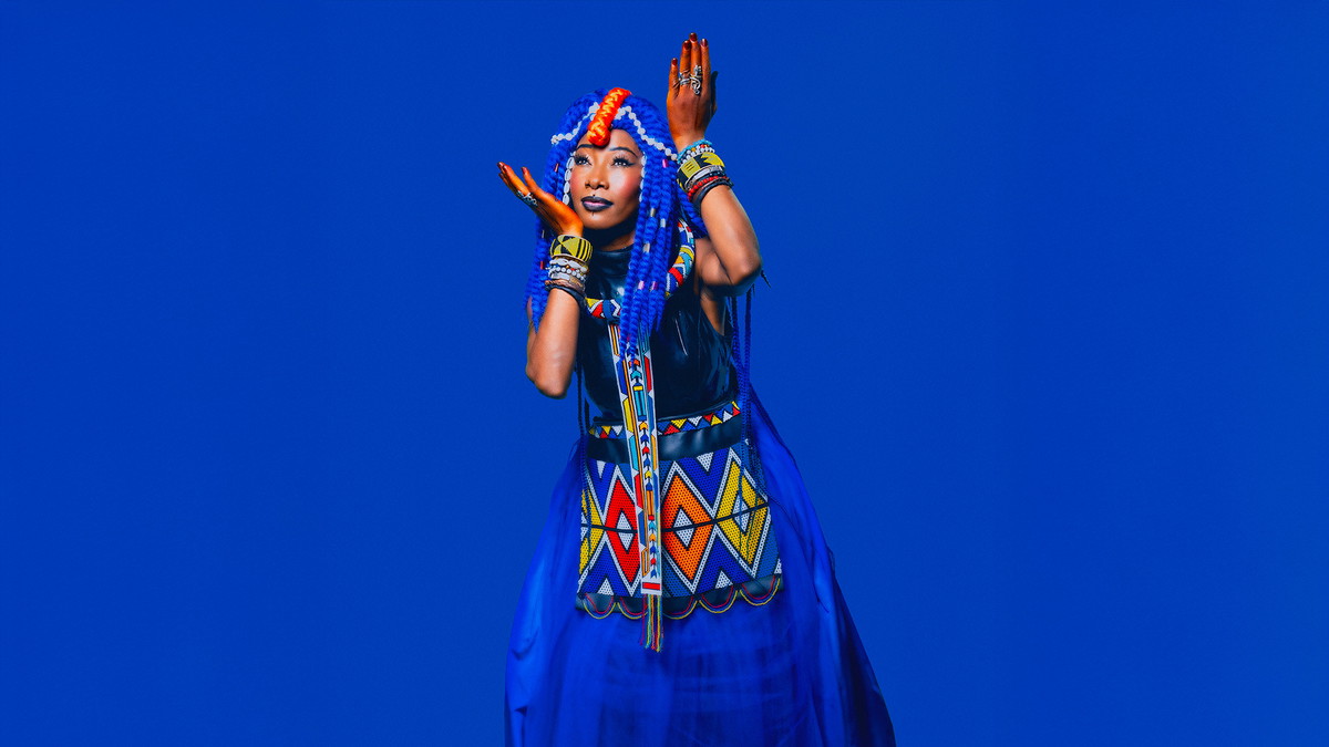Fatoumata Diawara steht in einem bunten Outfit in einer tanzenden Körperhaltung vor einer blauen Wand