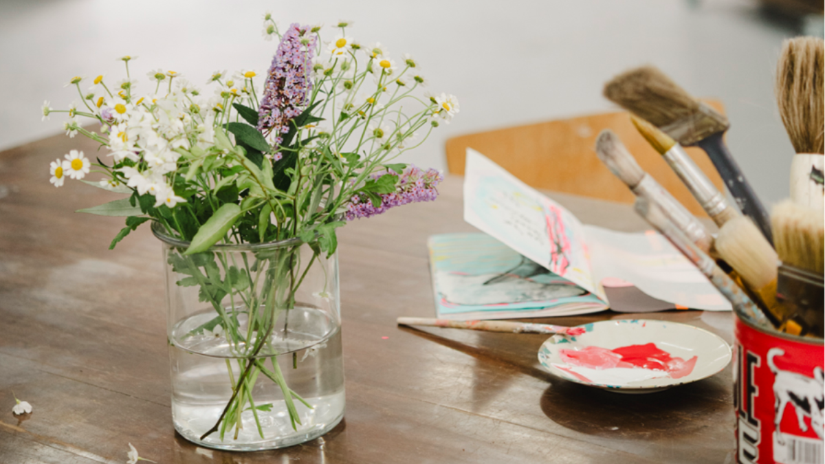 Tisch mit Glasvase und Frühlingsstrauch, Pinsel, Farbe und offenem Skizzenbuch