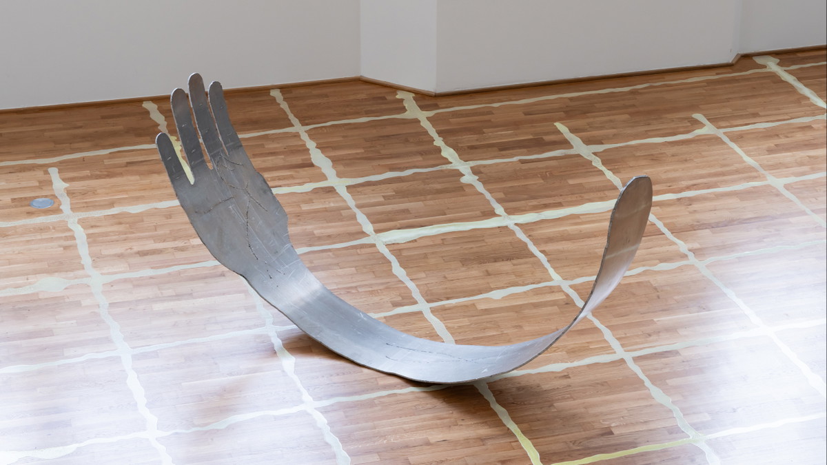 Eine Skulptur in Form eines Arms und einer Hand liegt auf Holzparkett; eine gitterartige Bodenzeichnung ist darunter.